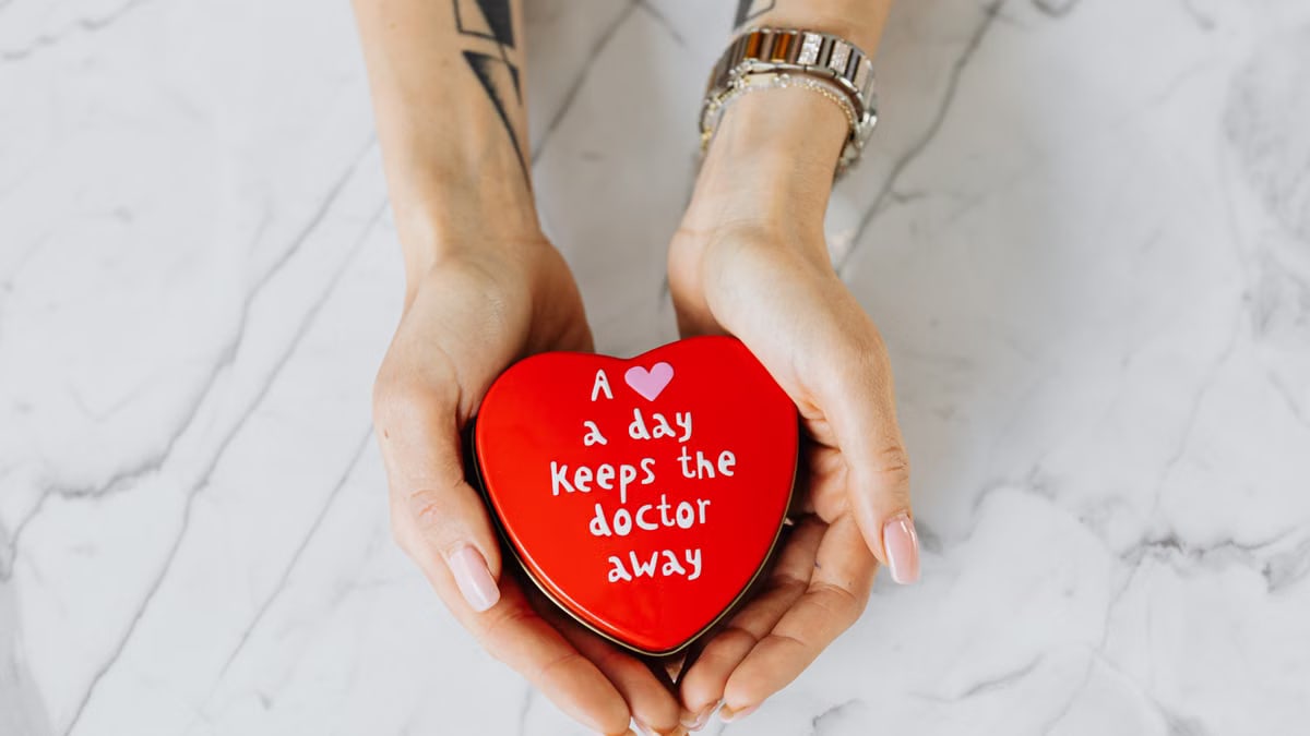 Hands holding a red heart-shaped tin with the message 'A heart a day keeps the doctor away' on a marble surface.